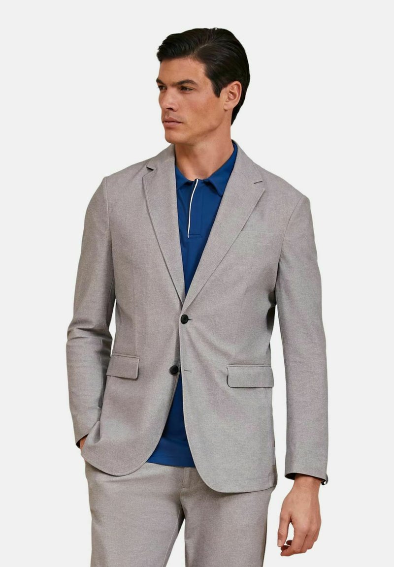 Blazer grigio chiaro su misura con rever a punta, due tasche frontali e bottoni neri, indossato sopra un polo blu con rifiniture bianche.