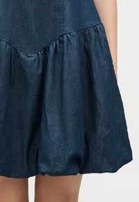 Σκούρο μπλε φούστα από τζιν με φούσκωμα στο τελείωμα που πέφτει πάνω από τα γόνατα, εμφανίζεται από το μέσο του μηρού έως τη μέση σε ανοιχτό φόντο.