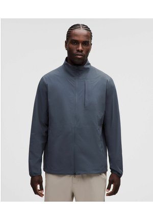 Man met gevlochten haar die een donkergrijze jas met rits en lichtbeige broek draagt tegen een effen lichte achtergrond.