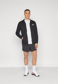 Black zip-up jacket with white logo, worn over a white t-shirt; paired with gray shorts and white athletic shoes. Plain background.