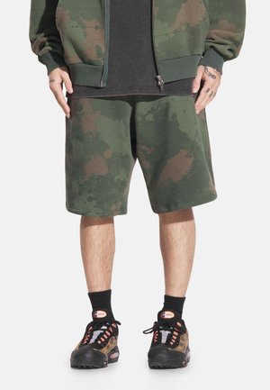 Zielone oliwkowe bawełniane szorty z wzorem chlapnięć w odcieniach brązu i jaśniejszych kolorach. Połączone z czarnymi trampkami i skarpetami. Widok z przodu.