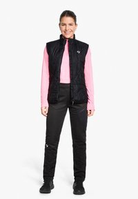 Black insulated vest over a pink long-sleeve shirt, paired with black pants featuring a textured design and athletic shoes, standing upright.