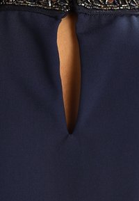 Tissu bleu marine avec un col en trou de serrure révélant la peau, accentué par une broderie complexe en perles noires et métalliques le long du bord.