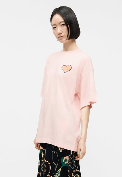 Γυναίκα με κοντά μαύρα μαλλιά που φοράει ένα φαρδύ παστέλ ροζ μπλουζάκι με γραφικό πορτοκαλί καρδιά, συνδυασμένο με μαύρη φούστα με αφηρημένα μοτίβα.