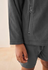 Grauer Fleece-Pullover mit Reißverschlusstasche, strukturiertem Stoff und langen Ärmeln, kombiniert mit passenden grauen Shorts aus glattem Material.