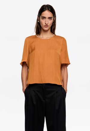 Blusa de manga corta y cuello redondo en un suave color mostaza con una textura ligera y fluida, combinada con pantalones negros de pierna ancha.