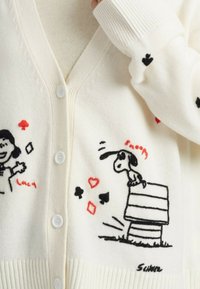 Weißer Cardigan mit schwarzen und roten gestickten Motiven von Snoopy auf seinem Hundebett und einem lächelnden Mädchen mit Spielkartensymbolen.