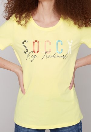 Żółta bawełniana koszulka z okrągłym dekoltem, ozdobiona czarną, czerwoną, różową, niebieską i białą typografią z napisem „SOCCX Reg. Trademark.”