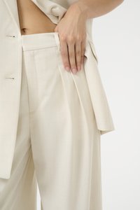 Blazer beige su misura con un solo bottone, abbinato a pantaloni a gamba larga con tasche laterali e una delicata texture del tessuto.