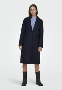 Lungo cappotto blu navy con rever a punta, chiusura a un bottone e tasche laterali. Texture liscia con un taglio dritto, indossato sopra una maglietta azzurro chiaro.