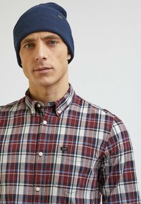 Granatowa czapka dzianinowa w rybkę i bordowa koszula w kratę z guzikami, z białymi i niebieskimi akcentami; posiada małe logo na lewej kieszeni piersiowej.