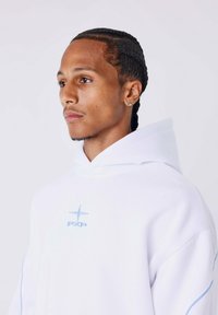 Man met gevlochten haar, draagt een diamantoorbel en een witte hoodie met lichtblauwe logo en piping, kijkt naar links tegen een effen achtergrond.