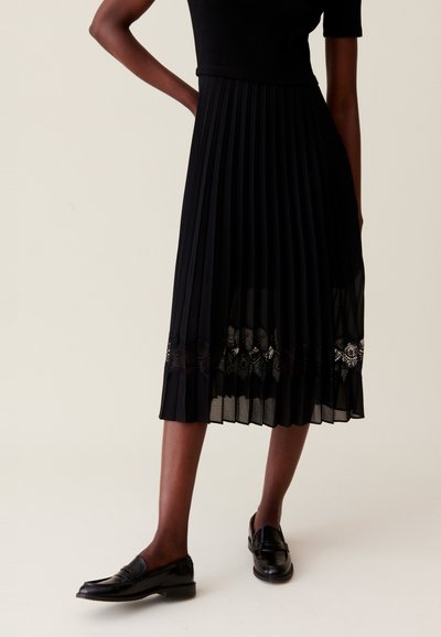 Zwarte geplooide rok met kantdetail aan de zoom, gecombineerd met glanzende zwarte loafers. De rok heeft een tailleriem en een vloeiende silhouet.
