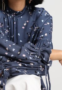 Nainen, joka pukee ylleen tummansinisen paidan, jossa on vaaleanpunaisia, mustia ja vaaleansinisiä abstrakteja pilkkuja sekä nyöritetyt hihat, jotka on solmittu nauhoilla.