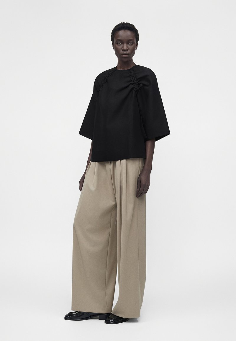 Model iført en sort oversized top med rynkede detaljer og brede beige bukser, stående mod en ensfarvet hvid baggrund.