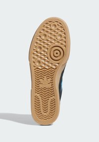 Čierna podrážka tenisky z textúrovaného gumového materiálu s kruhovými a trojuholníkovými vzormi a logom Adidas umiestneným v strede blízko päty.