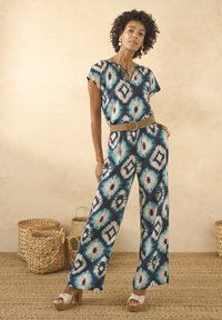 Giocoso tuta multicolore con un motivo tie-dye blu navy, verde acqua e crema, maniche corte e gambe ampie. Abbinata a una cintura beige e sandali con piattaforma.