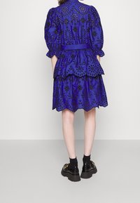 Robe bleue brodée avec manches bouffantes, design en couches, ourlet festonné et motifs floraux noirs. Associée à des chaussures à plateforme noires.