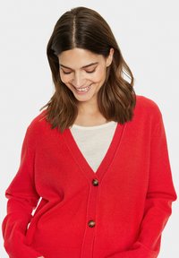 Röd kardiagan med V-hals, som har knappstängning och långa ärmar, tillverkad av ett mjukt, strukturerat tyg. Under finns en ljus färgad topp.