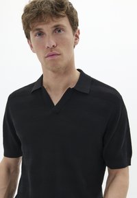 Črna polo majica s kratkimi rokavi z teksturiranim, prugastim dizajnom in ovratnikom v obliki črke V. Material izgleda mehak z rahlim sijajem.