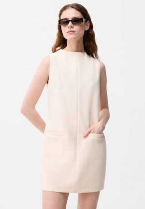 Femme portant une robe beige sans manches avec des poches avant et des lunettes de soleil rectangulaires foncées, debout sur un fond blanc uni.