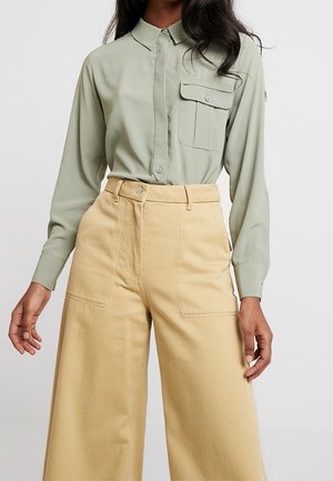 Kobieta w zielonym koszulowym swetrze z kieszenią na piersi i beżowych, szerokich spodniach z wysokim stanem na jednolitym tle.