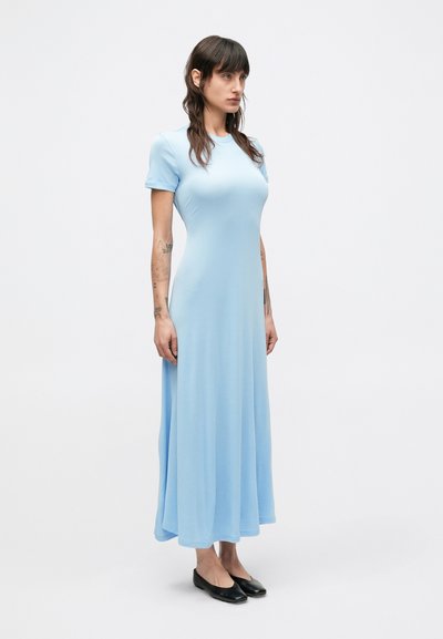 Svetlomodré maxi šaty s krátkym rukávom vyrobené z mäkkej látky. Plynulý dizajn s voľným strihom, siahajúce po členky. Nosené s čiernymi balerínkami.