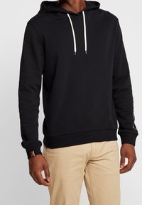 Sudadera con capucha de algodón negra con cordón en la capucha, puños y dobladillo acanalados, y detalles en blanco en las mangas, llevada sobre pantalones beige.