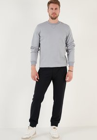 Felpa grigia con colletto a girocollo, polsini e orlo a coste, abbinata a pantaloni jogger neri. Il modello indossa sneaker bianche con accenti colorati.