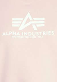 Felpa rosa chiaro con logo stampato bianco con lettere stilizzate "A" e "I" e testo che recita "ALPHA INDUSTRIES" e la posizione.