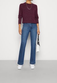 Pull en maille bordeaux, jeans évasés bleus, baskets blanches, collier chaîne argentée, tenant un petit sac à main blanc avec des accents noirs.