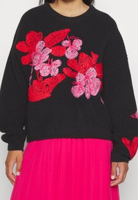 Svart stickad tröja med livfull röd och rosa blommig broderi. Har en rund hals och långa ärmar, ihopparad med en rosa veckad kjol.