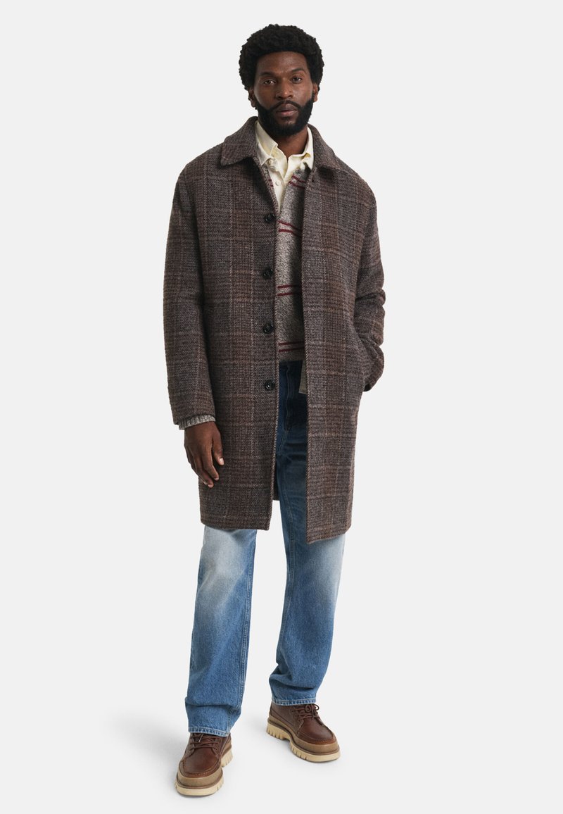 Man in een bruin geruit jasje, lichtgrijze trui met rode strepen, off-white shirt, blauwe jeans en bruine leren schoenen, staande met één hand in zijn zak.