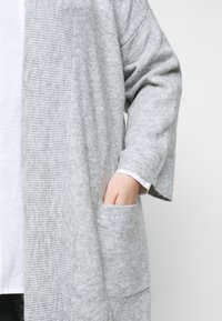 Cardigan tricoté gris clair à manches longues, texture côtelée et poche avec une main partiellement à l'intérieur sur une personne portant une chemise blanche.
