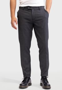 Homme portant un pantalon gris foncé bien ajusté et des chaussures en cuir noires à lacets, se tenant contre un fond clair.