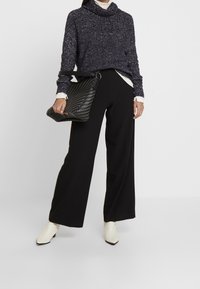 Zwarte wijde broek gecombineerd met een donker gestippeld truitje en crèmekleurige enkellaarsjes. Een gestructureerde zwarte handtas wordt in één hand vastgehouden.