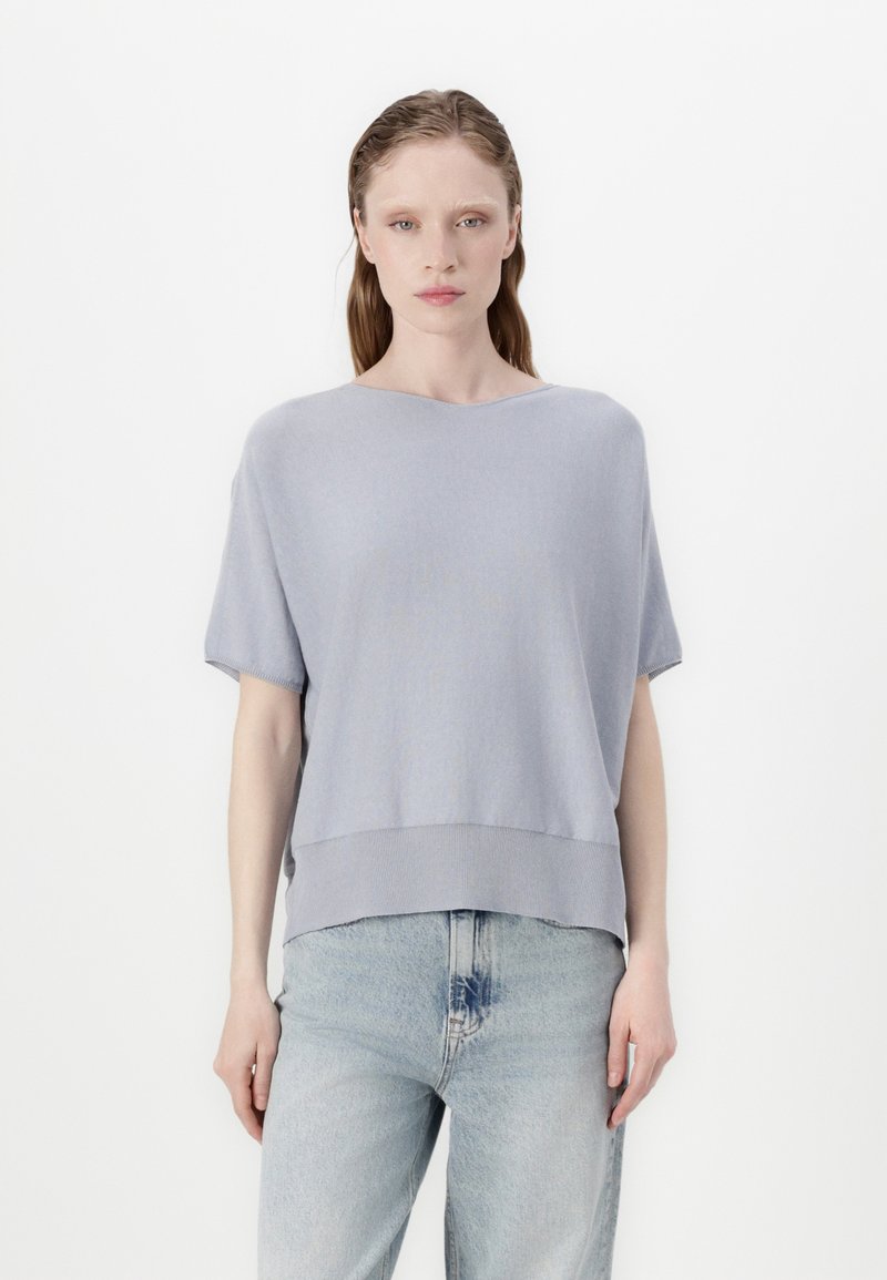 Lys grå strikket topp med korte ermer, vid halskant og ribbestrikket kant. Denne er kombinert med lyse denimjeans, og stoffet har en glatt tekstur.