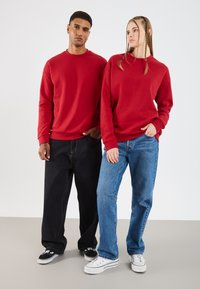 Rode sweatshirt met lange mouwen, gecombineerd met zwarte en blauwe jeans. De sweatshirts hebben ribbelboorden en -zoom en hebben een relaxte pasvorm.