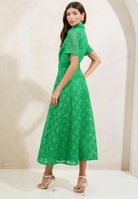 Abito verde in pizzo con colletto, maniche a palloncino corte e vita sagomata. Il tessuto presenta un motivo geometrico e si estende fino a metà polpaccio.