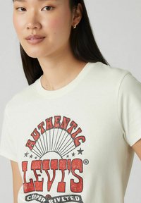 Camiseta de manga corta en color blanco roto con un atrevido estampado gráfico rojo que incluye la palabra "Levi's" y acentos de estrellas. Fabricada en suave algodón, con escote redondo.