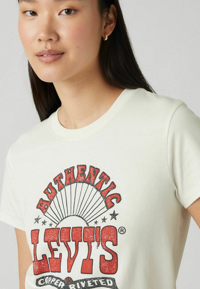 T-shirt à manches courtes couleur blanc cassé avec un imprimé graphique rouge audacieux portant le mot "Levi's" et des accents d'étoile. Tissu en coton doux, col rond.