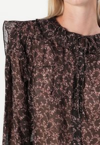 Blusa trasparente nera con un motivo floreale rosa, colletto arricciato, apertura con bottoni e tessuto testurizzato. Include arricciature sulle spalle e sui polsini.