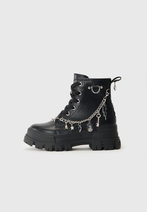 Schwarzer Leder-Stiefelette mit chunky Sohle, ausgestattet mit silbernen Kettenakzenten, Charms und einem runden Metallring. Strukturierte Oberfläche und Schnürdesign.