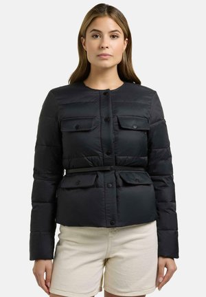 Kobieta w czarnej pikowanej kurtce z kieszeniami na guziki i cienkim paskiem, w połączeniu z jasnobeżowymi szortami na jednolitym tle.