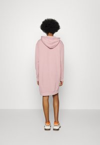 Rosa Kapuzensweatkleid, Midi-Länge mit langen Ärmeln, gerippten Bündchen und einer Kängurutasche. Getragen mit weißen Sneakers mit orangefarbenen Akzenten.