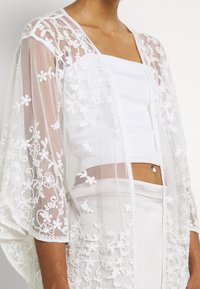 Kvindelig iført hvid blondeslør, blomsterkjole over en hvid crop top og lyse bukser, der viser maven og navlepiercing.
