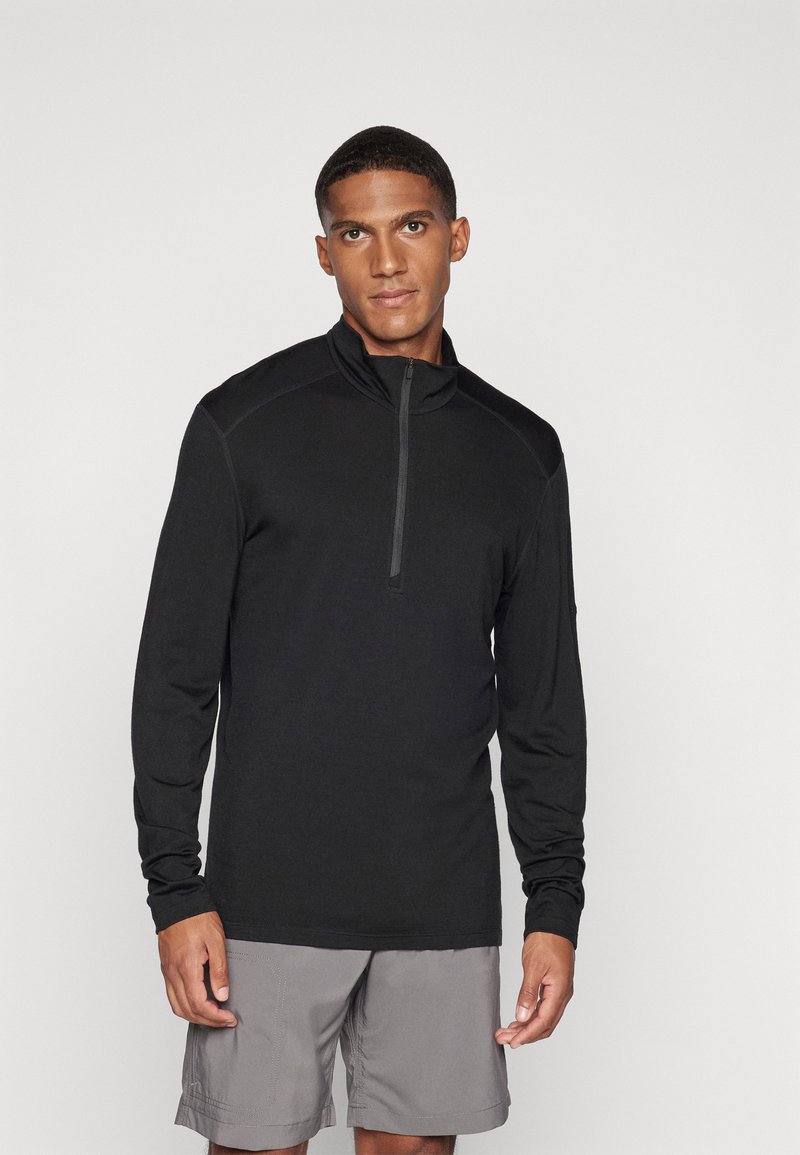 Pullover preto de mangas longas com gola de fecho de quarto, feito de material leve, combinado com calções cinzentos. Textura suave, design ajustado.