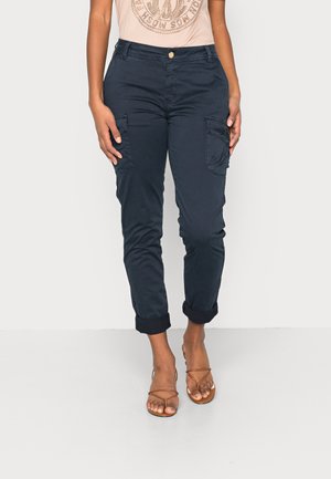 Frau trägt dunkelblaue Cargo-Hose mit hochgekrempeltem Saum, beige Grafik-T-Shirt und braune offene Sandalen mit Riemchen, steht auf hellem Hintergrund.