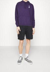 Lila hoodie med en framficka och logotyp, kombinerad med svarta cargoshorts och grå Vans-sneakers, kompletterad med vita ankelsockor.