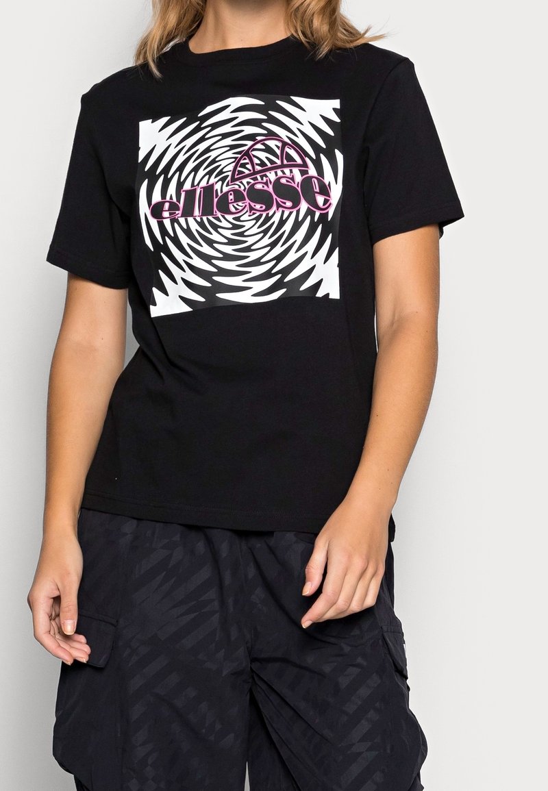 Personne portant un T-shirt noir avec un graphique en spirale blanc et noir et un logo rose "ellesse", associé à un pantalon noir à motifs.